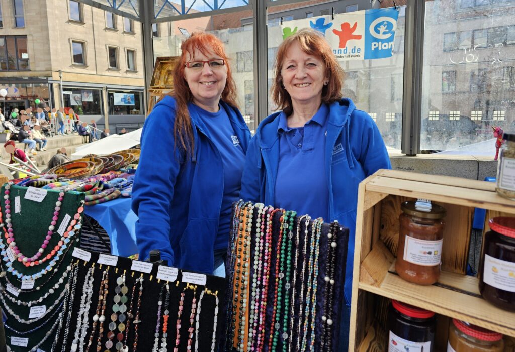 Two smiling women in blue jackets stand behind a market stall displaying colorful beaded necklaces, bracelets, and jars of homemade goods. People and buildings are visible in the background.