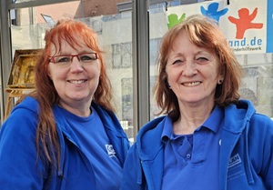 Two women with reddish hair, wearing blue shirts and blue zip-up hoodies, stand indoors smiling. There is a window behind them with colorful people-shaped figures and part of a German website name visible.