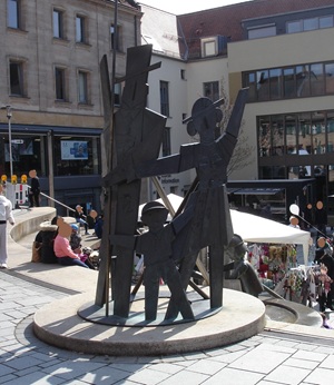 A metal sculpture depicting three abstract, human-like figures of varying heights stands on a circular base in a busy urban square with market stalls and people in the background.