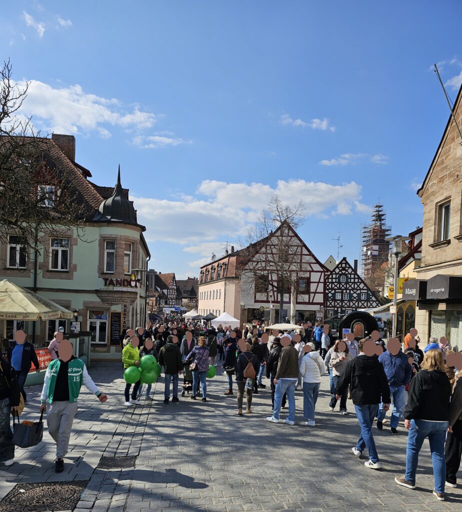 A lively street scene in a European town shows many people walking and talking under a sunny sky. Historic buildings line the street, with shops, cafes, and a group holding green balloons.
