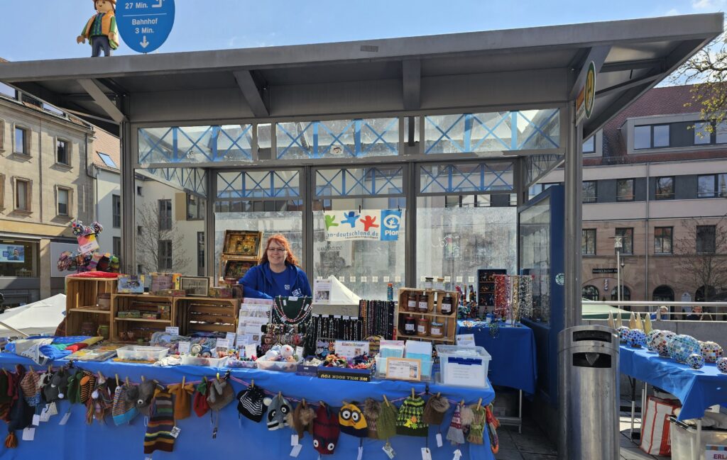 A woman stands behind a market stall covered with blue cloth, displaying handmade crafts, jewelry, hats, and stuffed animals in an outdoor urban setting with buildings and a glass shelter in the background.