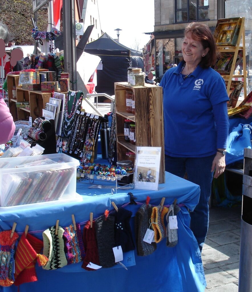 A woman in a blue shirt stands smiling behind a table at an outdoor market. The table displays colorful handmade items, including knit mittens hanging on a clothesline and various crafts arranged neatly.