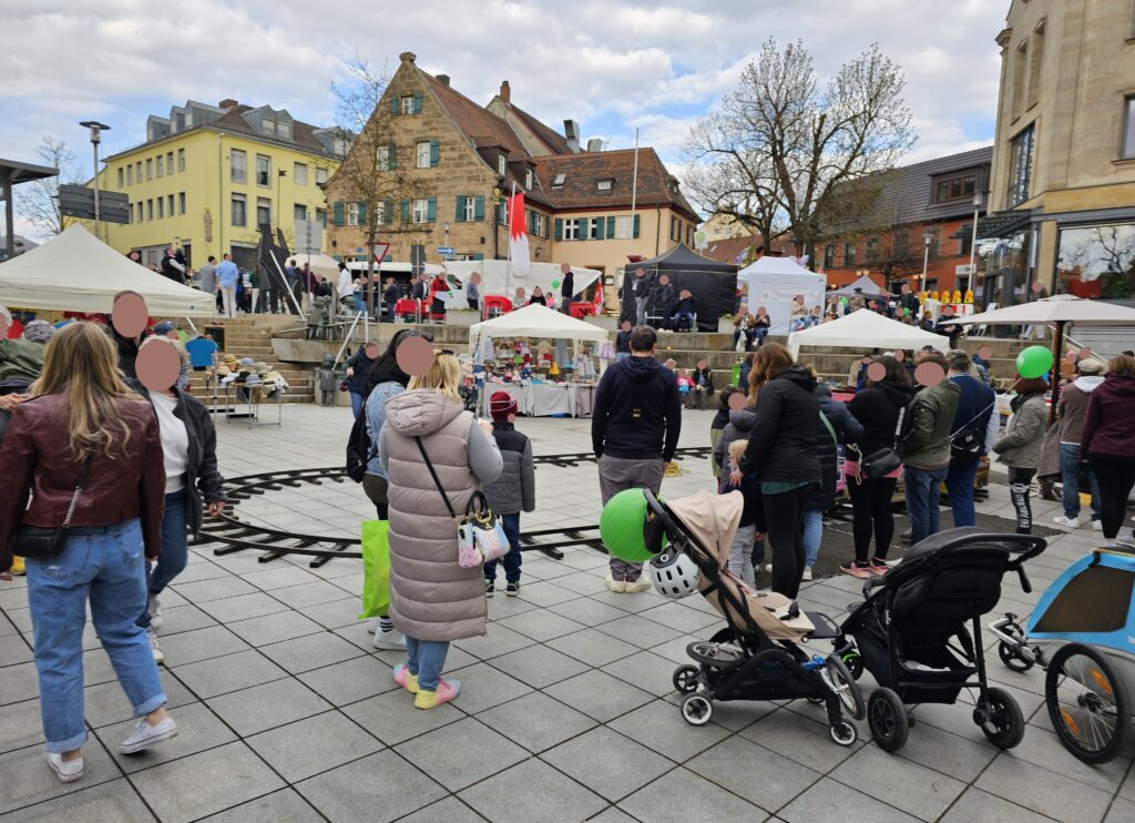 A crowd of people, including families with children and strollers, gather in a town square with tents and stalls. A small rideable train track circles the center, and a stage with musicians is visible in the background.