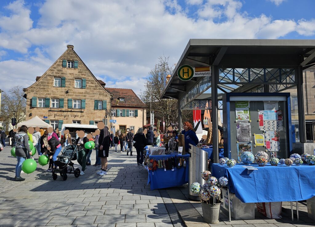 A lively outdoor market scene with people walking, some holding green balloons. Stalls display goods, including a table with colorful decorated balls. A stone building and a kiosk covered with posters are visible under a partly cloudy sky.