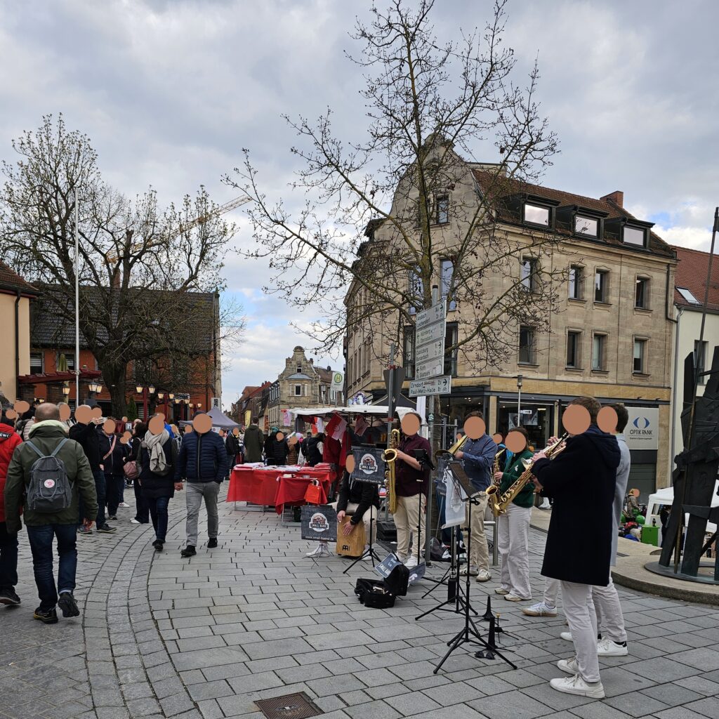 A street band performs for a crowd in a European town square. People gather around, some watching, while market stalls and old buildings line the street under a cloudy sky.