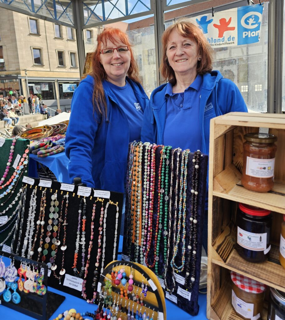 Two smiling women in blue tops stand behind a display of colorful beaded necklaces and handmade goods at a market stall. Jars and wooden shelves are also visible on the right side. A busy street scene is in the background.
