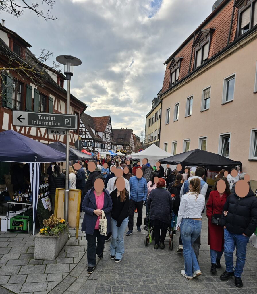 A busy outdoor street market with many people walking between vendor stalls. Traditional timber-framed buildings and a sign for tourist information are visible under a cloudy sky.