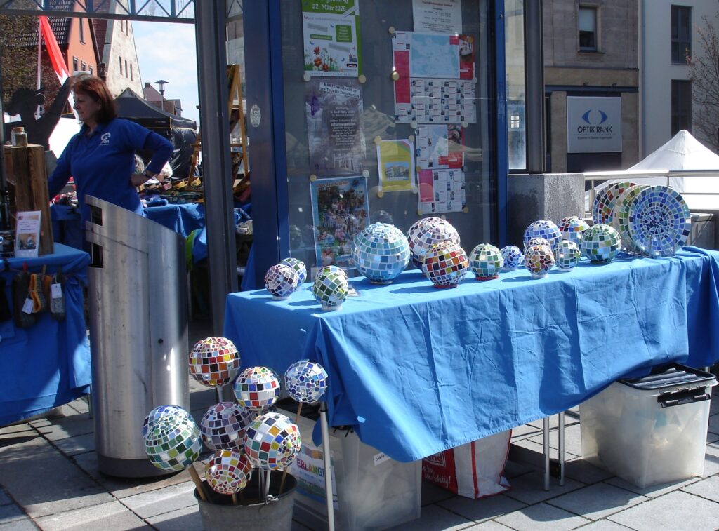 A table with a blue cloth displays various mosaic glass spheres in different sizes. A woman stands nearby, and informational posters are visible behind the table in an outdoor market setting.