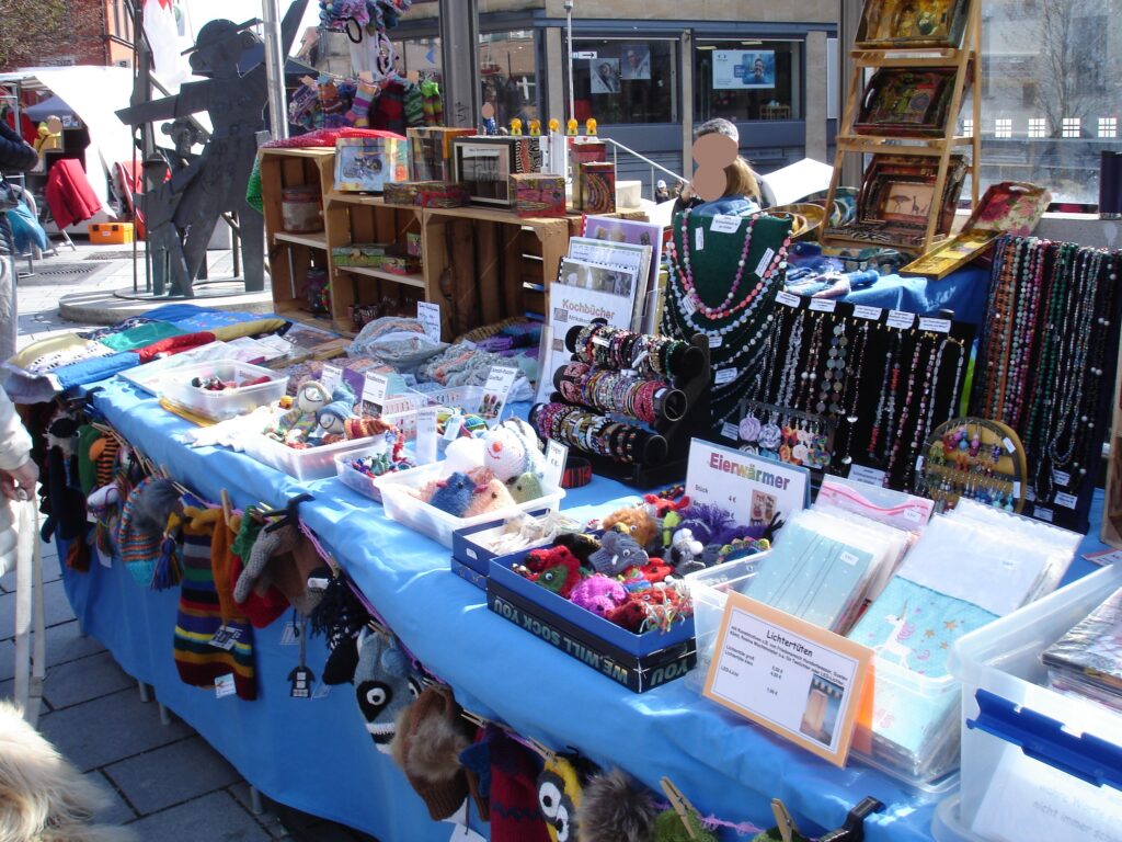 A colorful outdoor market stall displays handmade crafts, jewelry, knitted items, small toys, and accessories on a table covered with a blue cloth. Wooden shelves and racks hold more items behind the display.