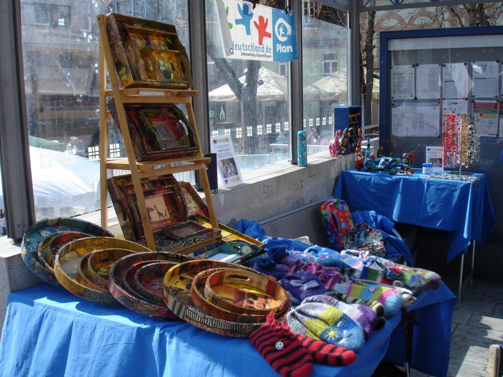 A colorful market stall displays handmade crafts, including felted mittens, patchwork textiles, trays, and beaded jewelry, all arranged on blue tablecloths under natural light. Posters and flyers are visible in the background.