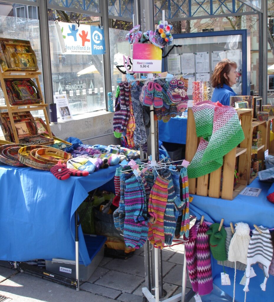 A market stall displays colorful knitted socks, hats, mittens, and scarves on racks and tables covered with blue cloth. Wooden boxes hold more knitwear. A woman stands behind the stall, and posters are visible in the background.