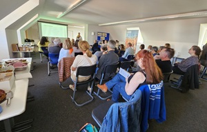 A group of people seated in a bright conference room, attentively listening to a presenter at the front near a large projected screen, with refreshments on a table nearby.