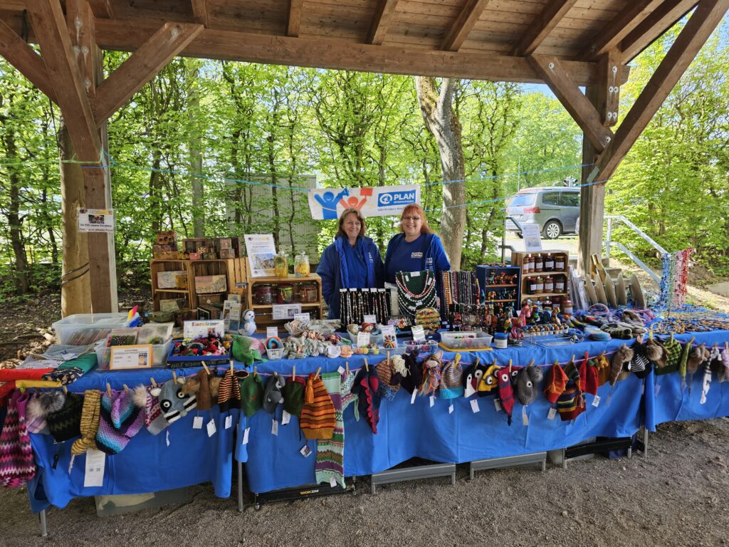 Two people stand behind a market stall under a wooden shelter, selling colorful handmade crafts, jewelry, and knitted items. The table is covered with a blue cloth and surrounded by trees and greenery in the background.
