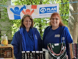 Two women in blue jackets stand behind a table displaying beaded jewelry at an outdoor stall. A “PLAN International” banner hangs above them, and green trees can be seen in the background.