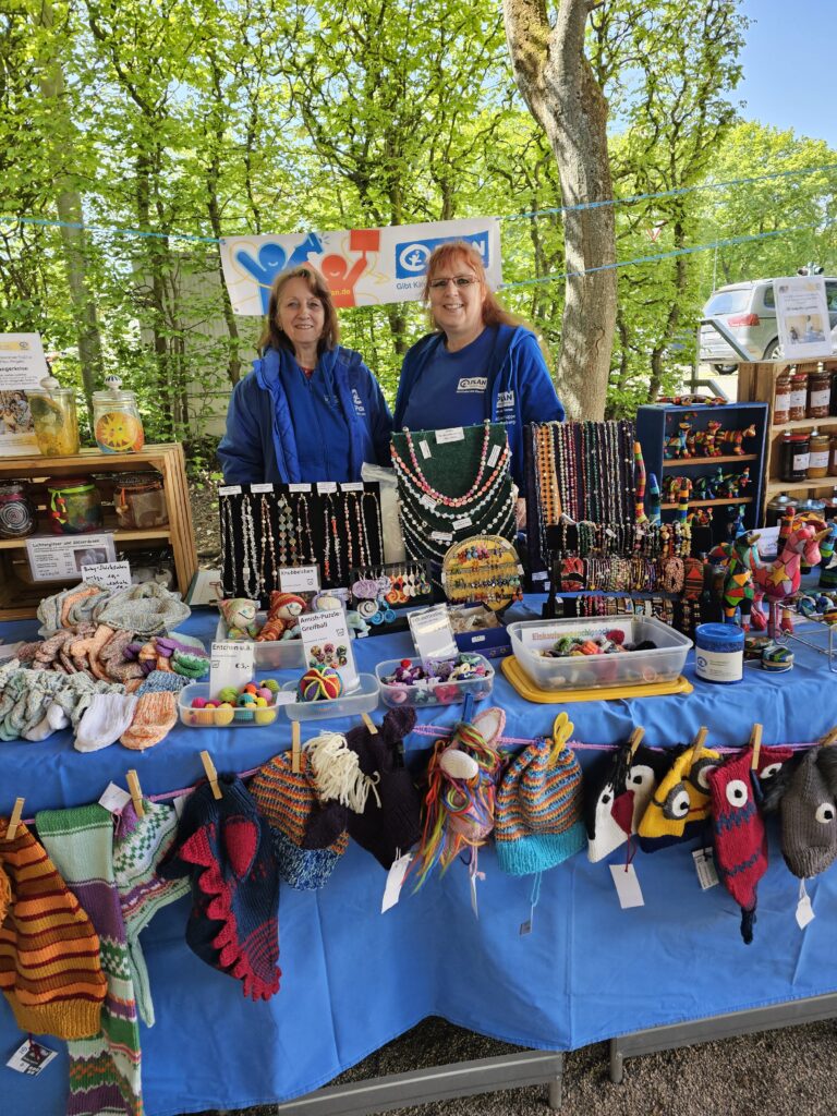 Two women in blue jackets stand behind a colorful outdoor market stall displaying handmade crafts, knitwear, jewelry, stuffed animals, jams, and honey jars on a blue tablecloth with trees in the background.