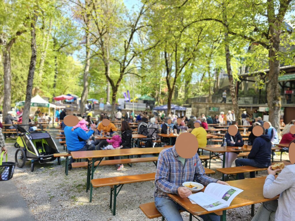 People sit at wooden tables in an outdoor park café under leafy green trees. Some are eating or reading, while others talk. Strollers and bikes are nearby, and more people gather around stalls in the background. Faces are blurred.