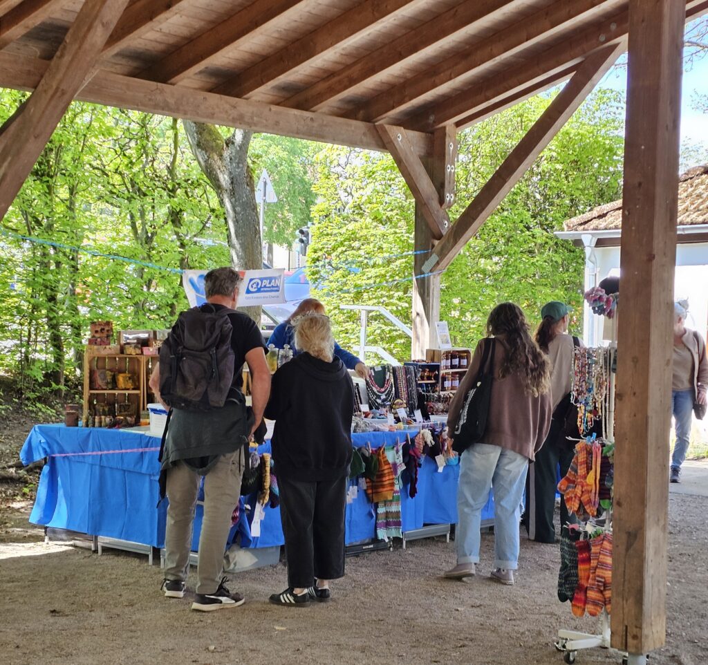 Four people stand under a wooden pavilion, browsing colorful handmade crafts and jewelry displayed on tables covered with blue cloths at an outdoor market surrounded by trees.