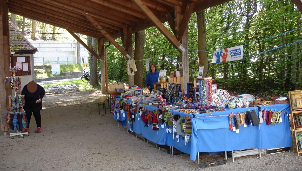 A covered outdoor market stand displays colorful handmade crafts, including hats, scarves, toys, and jewelry on blue tablecloths. One person stands behind the stall, another browses nearby. Trees are in the background.
