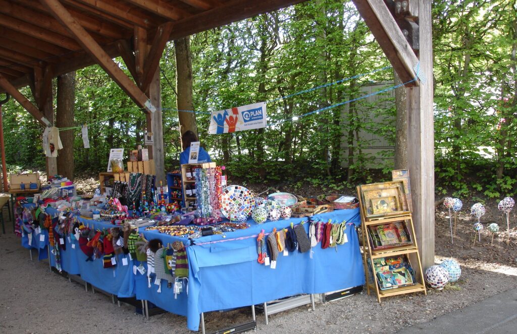 A covered outdoor market stall with a blue tablecloth displays colorful handmade crafts, including knitted items, jewelry, and framed pictures, with trees and greenery in the background.