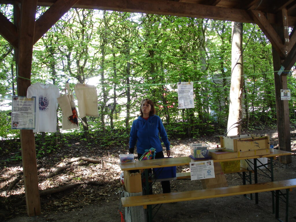 A woman stands under a wooden shelter in a forested area, looking up. T-shirts and papers hang on a line, with tables holding boxes and supplies in the foreground. Sunlight filters through the green trees.