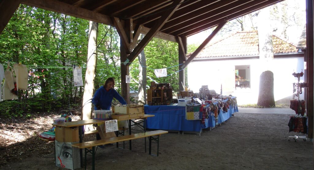 A person arranges items at a market stall under a wooden pavilion. The tables are covered with blue cloths displaying various goods. Trees and a white building are visible in the background.