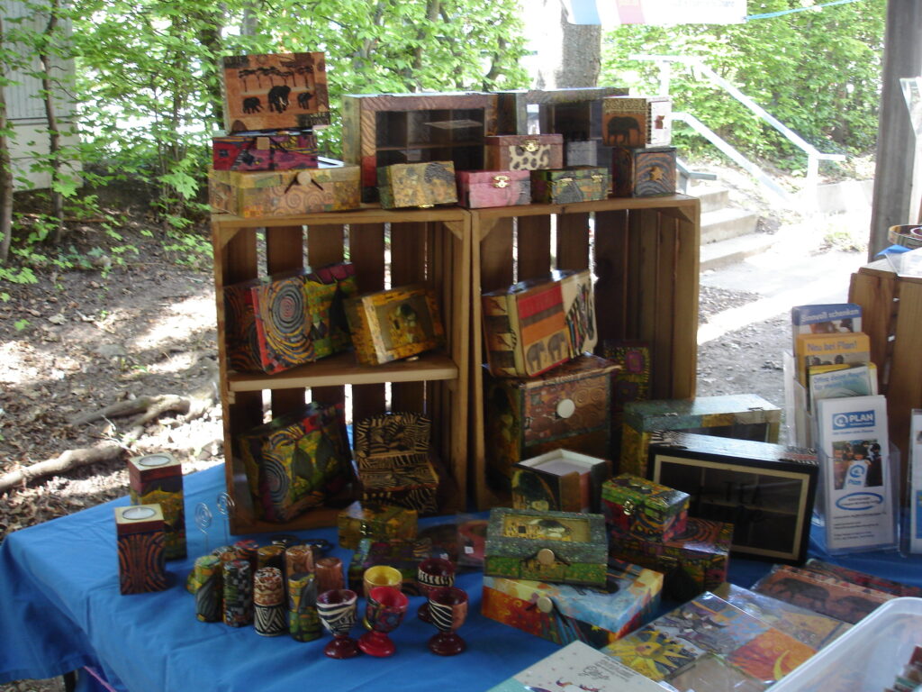 Hand-painted wooden boxes, bowls, and crafts are displayed on wooden crates and a blue tablecloth at an outdoor market stall surrounded by trees. Informational pamphlets are stacked on the right side of the table.