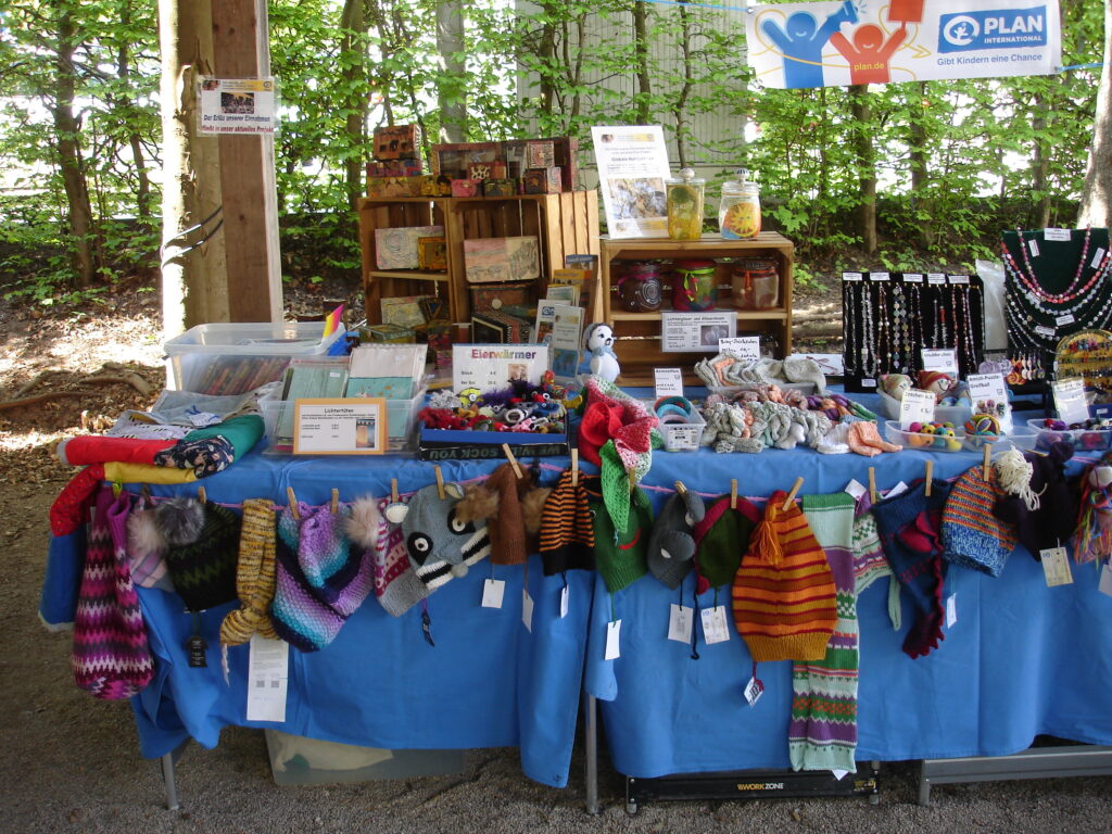 A market stall displays colorful knitted mittens, hats, and plush toys on a blue tablecloth, with jewelry, cards, and small crafts on shelves behind, set outdoors with trees in the background.