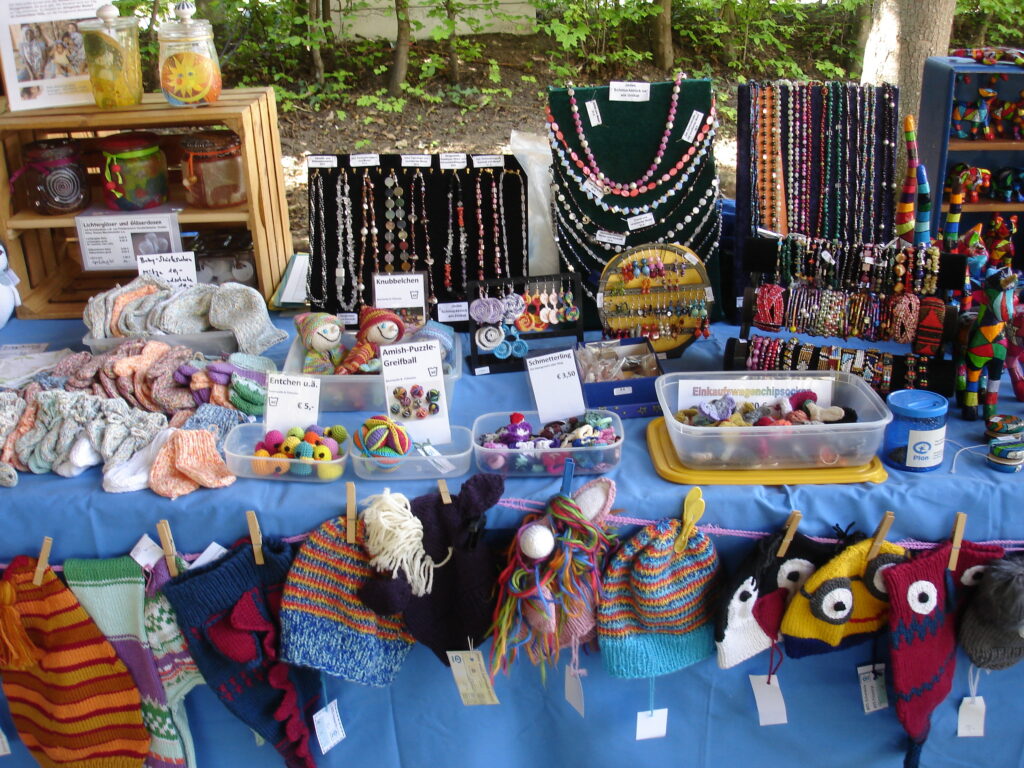 A market stall displays colorful handmade items, including knitted hats, socks, beaded necklaces, bracelets, and small crafts, all arranged on a blue tablecloth with some items hanging in front.