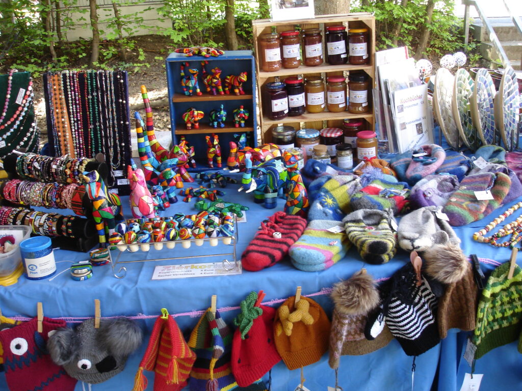 A market stall displays colorful knit hats, mittens, beaded jewelry, jars of preserves, fabric crafts, and toys on a blue tablecloth, with trees and greenery in the background.