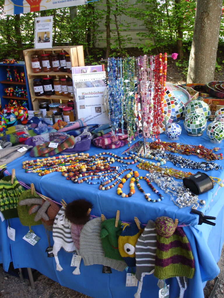 A market stall displays colorful handmade bead jewelry, knitted hats with animal designs, jars of preserves, felt items, and mosaic decorations, all arranged on a blue tablecloth outdoors among trees.