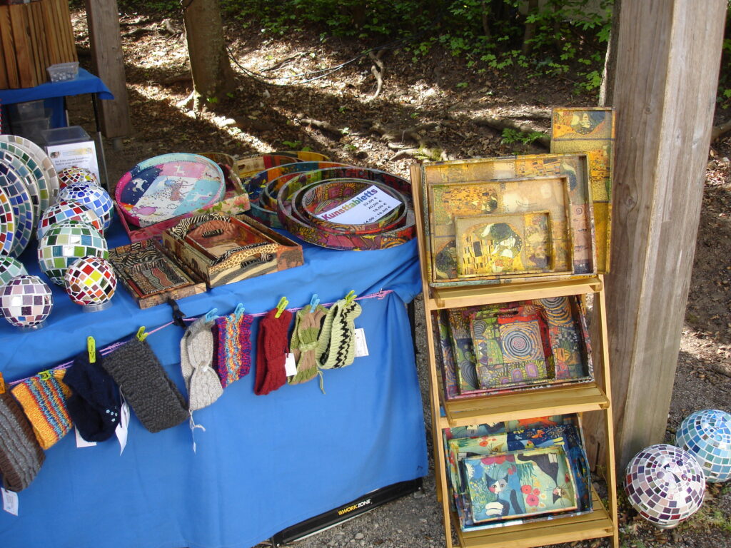 A market stall displays colorful mosaic plates, patterned trays, knitted headbands, and hand-painted wooden items on a blue table and wooden shelves, surrounded by trees and dappled sunlight.