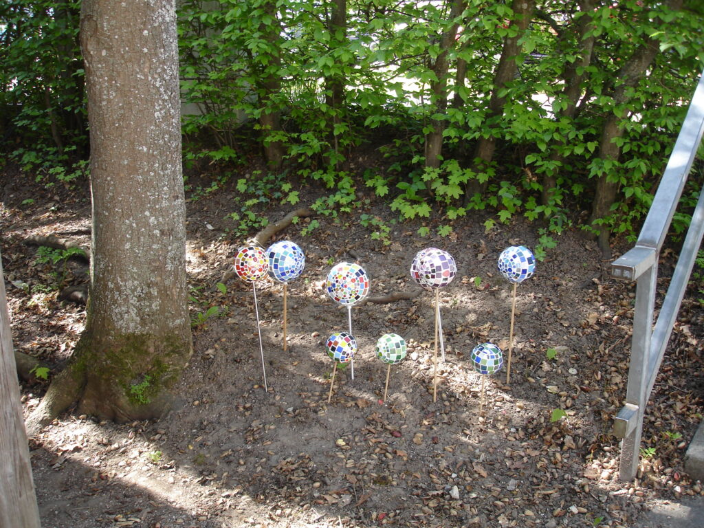 Eight decorative mosaic spheres on sticks are arranged in a patch of dirt near a tree and leafy bushes. The spheres are covered with colorful tiles and vary in size, standing in sunlight and shade.