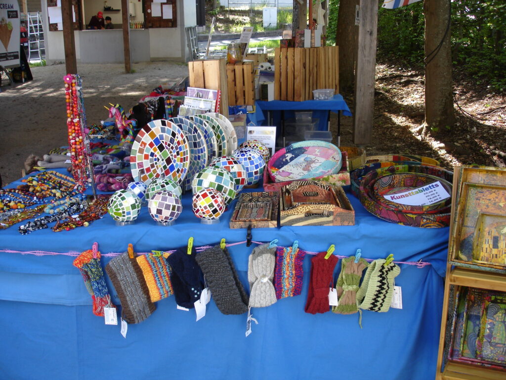 A market stall with a blue tablecloth displays colorful mosaic plates and balls, beaded necklaces, knitted items, tapestries, and framed artwork, set outdoors under a wooden shelter.