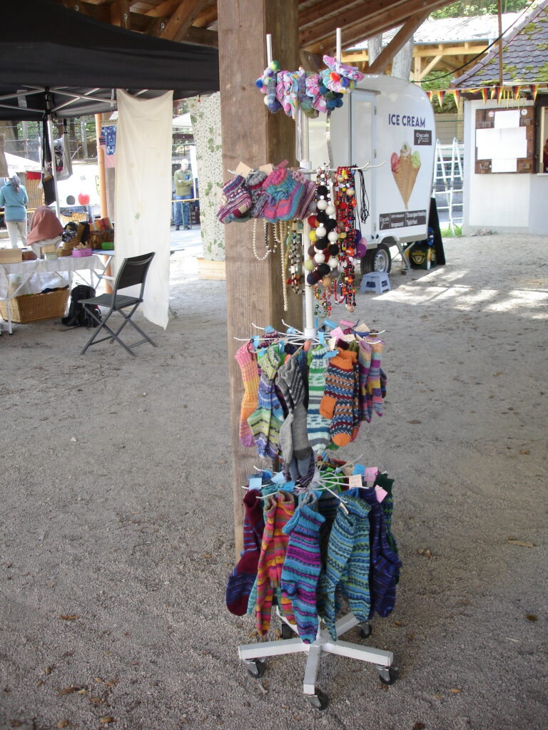 A display stand at an outdoor market shows colorful knitted socks, mittens, and small crocheted items. Market stalls, a trailer with an Ice Cream sign, and chairs are visible in the background.