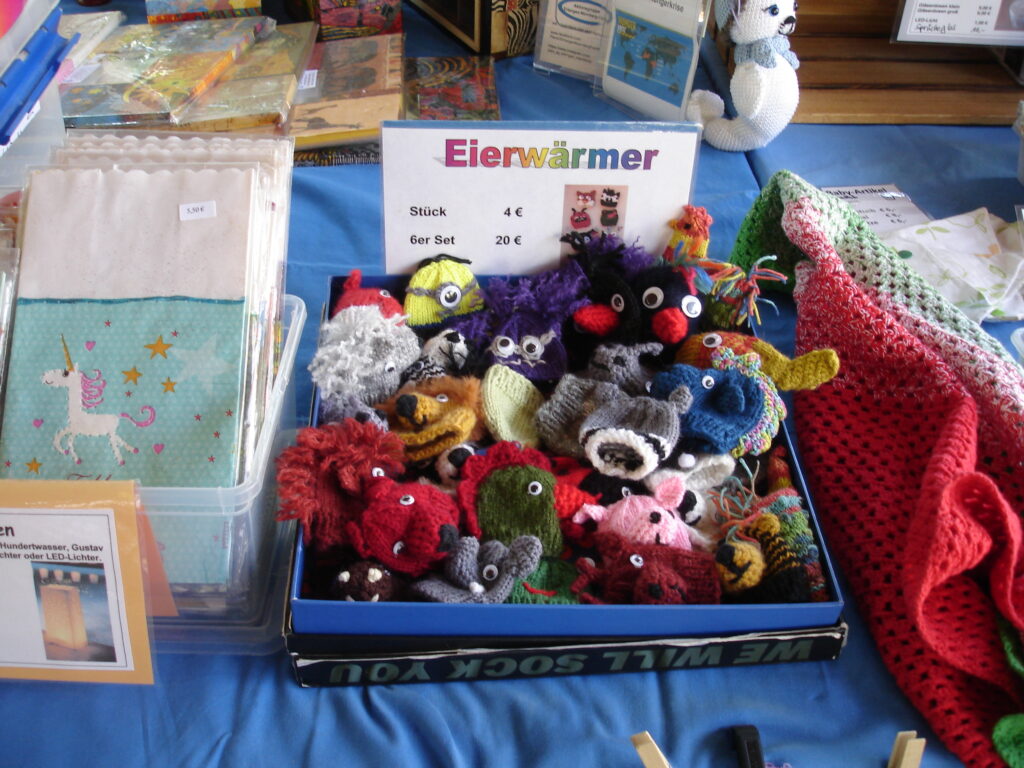 A box labeled Eierwärmer displays colorful, knitted egg cozies shaped like animals with googly eyes. The table also shows crocheted items, napkins, and other handmade crafts on a blue cloth.