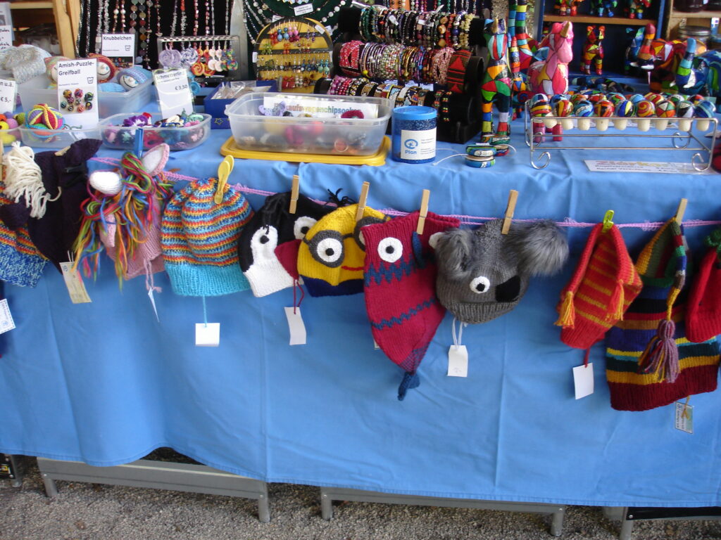 Colorful knitted hats with playful designs and faces hang on a clothesline above a blue table, with various beaded jewelry and craft supplies displayed behind them at a market stall.