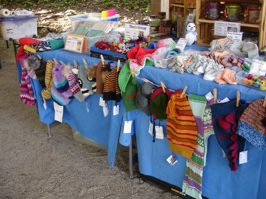 A market stall displays colorful hand-knitted hats, scarves, and mittens hanging on a blue cloth, with more handmade items and crafts arranged on tables in the background.