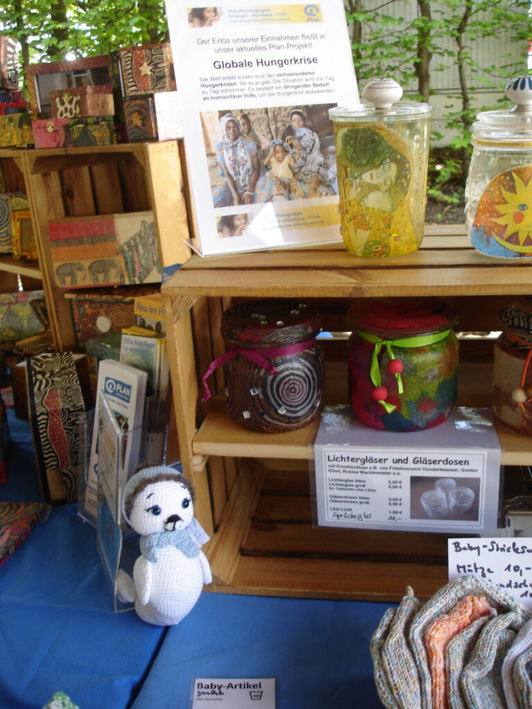 A market stall displays colorful handmade crafts, jars, and a crocheted penguin toy. Wooden crates hold products, and a sign about fighting global hunger is visible in the background. A blue cloth covers the table.