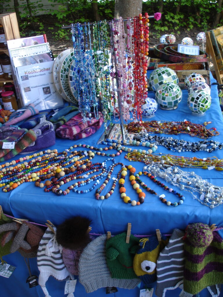 A market stall displays colorful beaded necklaces, bracelets, and knitted hats on a blue tablecloth. Glass mosaic lanterns are in the background, and a few felted accessories are arranged on the table.
