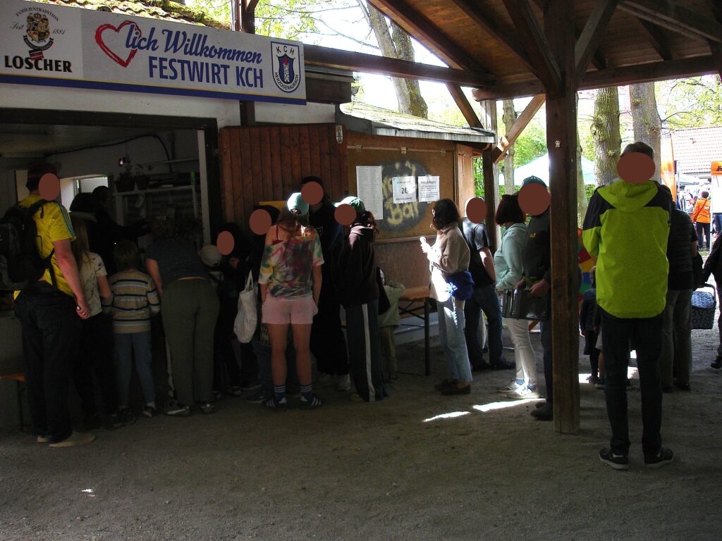 A group of people, many with backpacks, stand in line outside a wooden food stand under a shaded shelter. A sign above reads “Herzlich Willkommen Festwirt KCH.” Trees and sunlight are visible in the background.