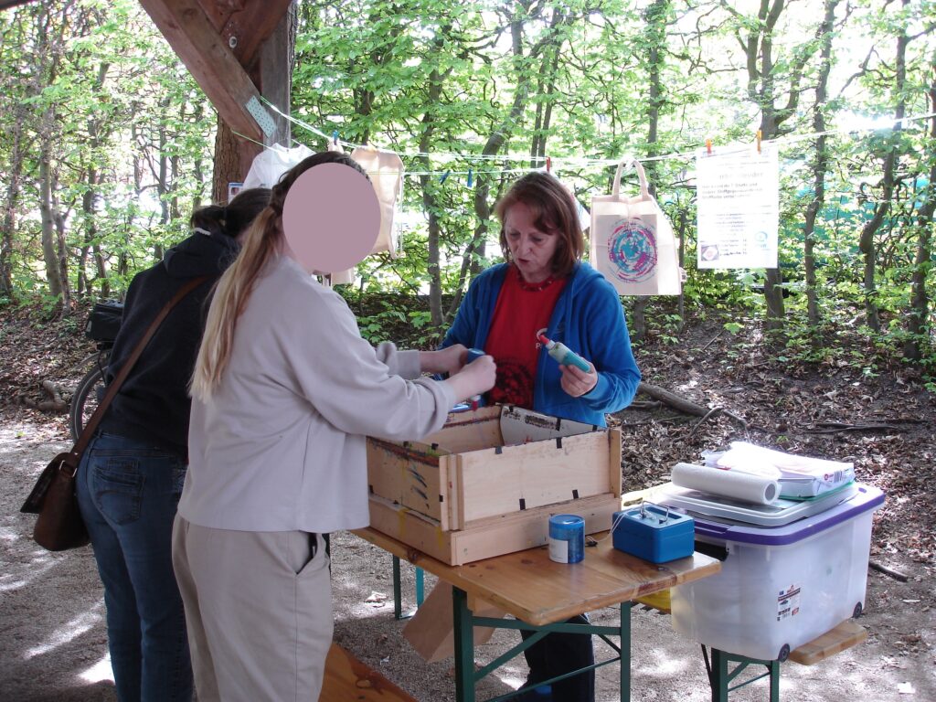 Two people stand at a wooden table outdoors, interacting with a woman in a blue jacket who is holding an item. There are boxes, papers, and bags on the table, and trees are visible in the background.