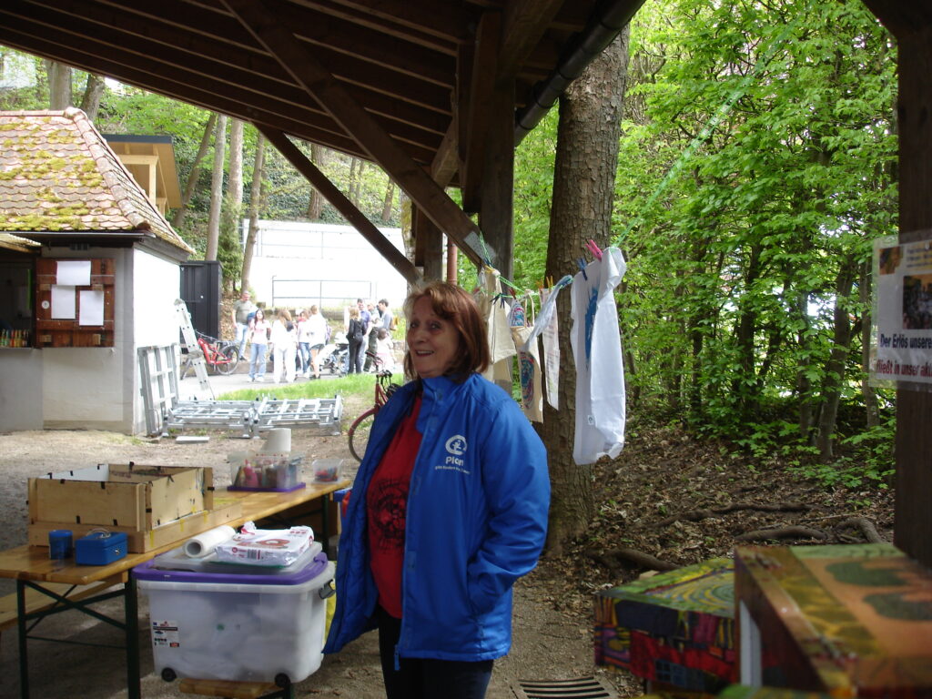 A woman in a blue jacket stands smiling under a wooden shelter at an outdoor event, with tables displaying items and a group of people gathered in the background near trees and greenery.
