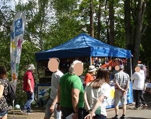 Auf einer Messe oder einem Markt im Freien gehen Menschen an einem blauen Zelt vorbei, das von Bäumen umgeben ist. In dem Zelt werden verschiedene Artikel zum Verkauf angeboten, und in der Nähe steht eine vertikale Fahne mit bunten Blumenmustern. Die Gesichter sind zum Schutz der Privatsphäre unscharf.