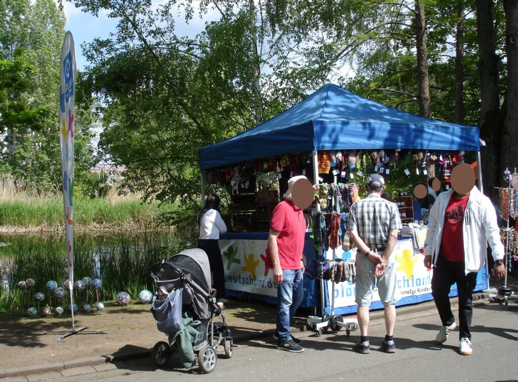 Ein Marktstand mit blauem Vordach in einem Park verkauft verschiedene Kunsthandwerksprodukte, während drei Erwachsene und ein Kinderwagen in der Nähe stöbern. An einem sonnigen Tag sind im Hintergrund Bäume und ein Teich zu sehen.