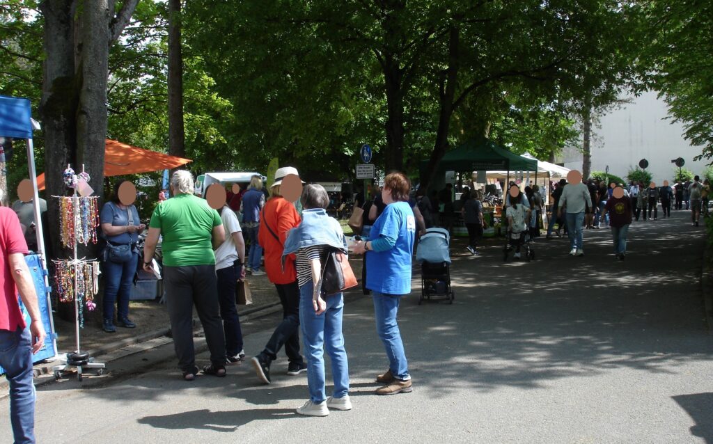 Auf einem Straßenmarkt oder einer Messe versammeln sich die Menschen im Freien und stehen und gehen zwischen überdachten Ständen. Bäume spenden Schatten, und es werden verschiedene Artikel zum Verkauf angeboten. Die Atmosphäre wirkt ungezwungen und lebendig.