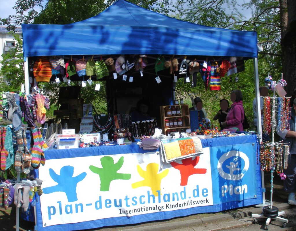 Ein Marktstand in einem blauen Zelt bietet bunte Strickmützen, Socken und handgefertigten Schmuck an. Auf einem großen Schild steht plan-deutschland.de Internationales Kinderhilfswerk. Mehrere Menschen stehen hinter und neben dem Stand.