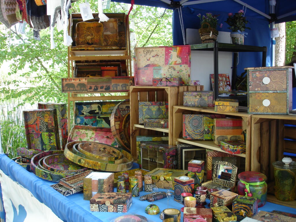 An einem farbenfrohen Marktstand im Freien werden verschiedene handbemalte Holzkisten, Tabletts und Dekorationsartikel mit lebhaften Mustern und Designs ausgestellt, umgeben von Grünpflanzen und Bäumen.