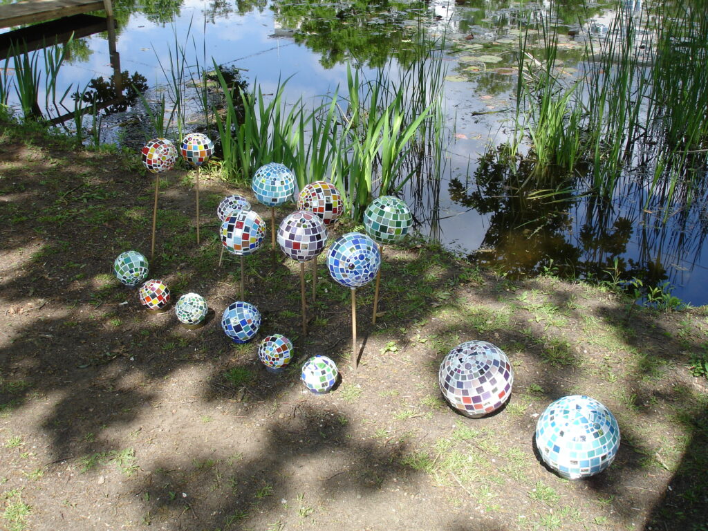 Mehrere bunte Mosaikkugeln unterschiedlicher Größe sind auf und über dem Boden neben einem Teich angeordnet, einige auf Stöcken zwischen grünen Pflanzen und Schilf, wobei sich Bäume und Himmel im Wasser spiegeln.