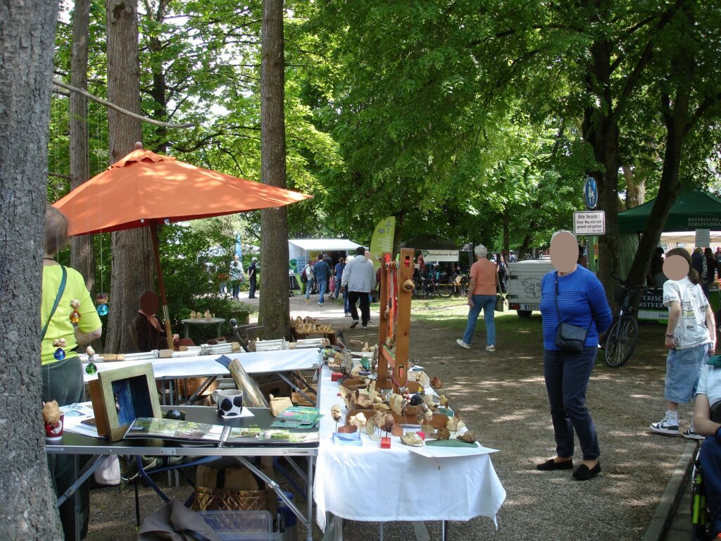 Auf einem Markt unter freiem Himmel stöbern die Menschen an verschiedenen Tischen mit Kunsthandwerk und Kunstwerken unter Bäumen. Einige gehen spazieren, andere begutachten die Waren. Ein orangefarbener Sonnenschirm beschattet einen Tisch, und die Umgebung ist von Grün und Sonnenlicht erfüllt. Die Gesichter sind verschwommen.