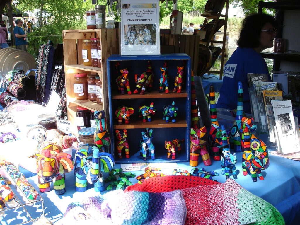 An einem Marktstand werden bunte, handgefertigte Tierfiguren und gehäkelte Gegenstände auf einem Tisch ausgestellt, wobei Holzkisten mit Einmachgläsern und eine Person hinter der Auslage stehen. Das Sonnenlicht fällt durch die Bäume im Hintergrund.
